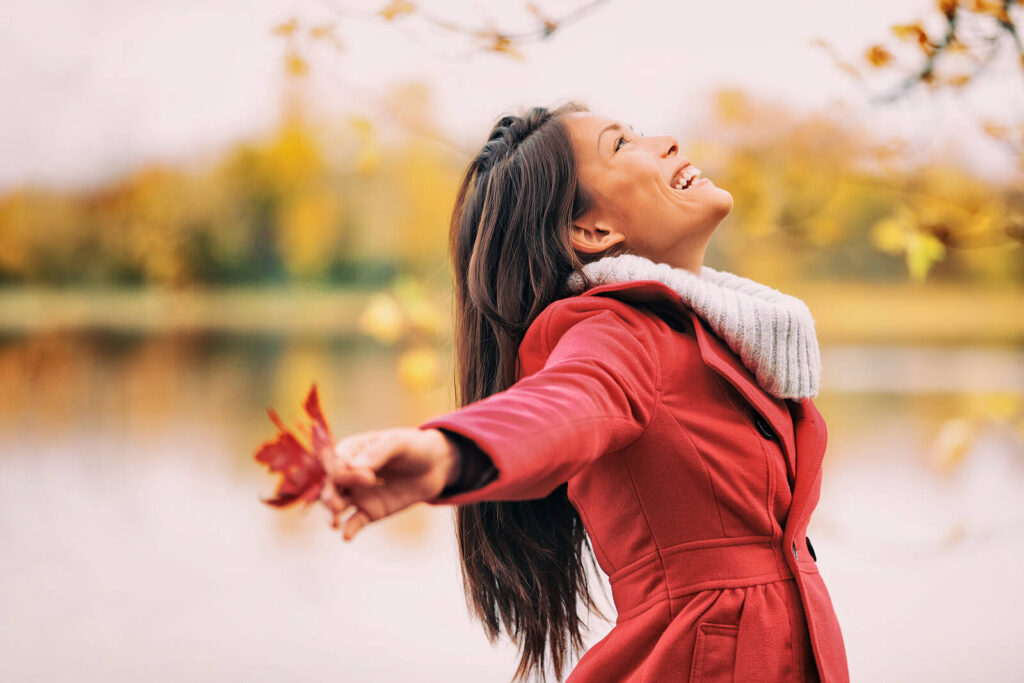 Image of a smiling woman standing outside smiling up at the sky while holding leaves. Struggling with an eating disorder can be overwhelming. With the help of eating disorder recovery coaching in the United Kingdom you don't have to face it alone.