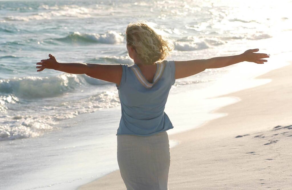 Image of a happy woman standing on the beach with her arms out. Create a healthier relationship with food and yourself with the support of Healthy Self Recovery and a skilled eating disorder recovery coach in the UK.