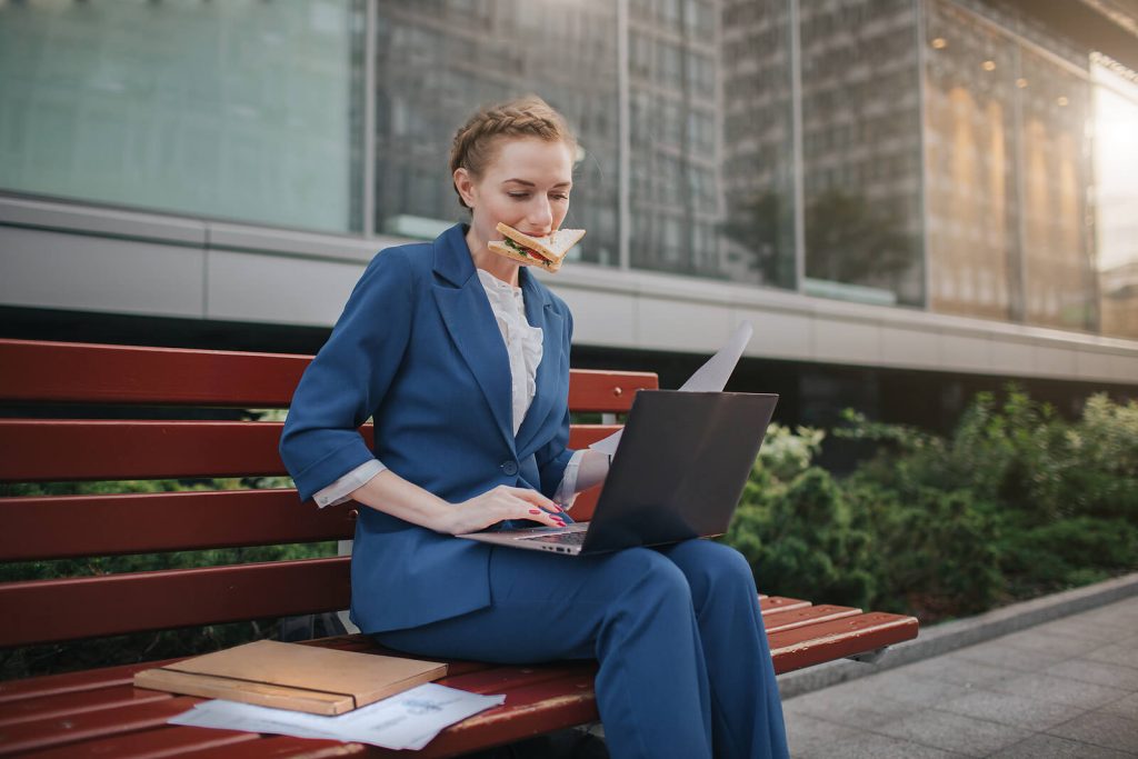 stressed woman on bench | eating disorder recovery coaching in the uk | eating disorder recovery coach in the uk | eating disorder coaching | british eating disorder recovery coach | England | Scotland | Ireland Image of a stressed professional woman sitting on a bench outside eating a sandwich while typing on a laptop. With a British eating disorder recovery coach, you can begin to manage your eating disorder in healthy ways as a busy professional.