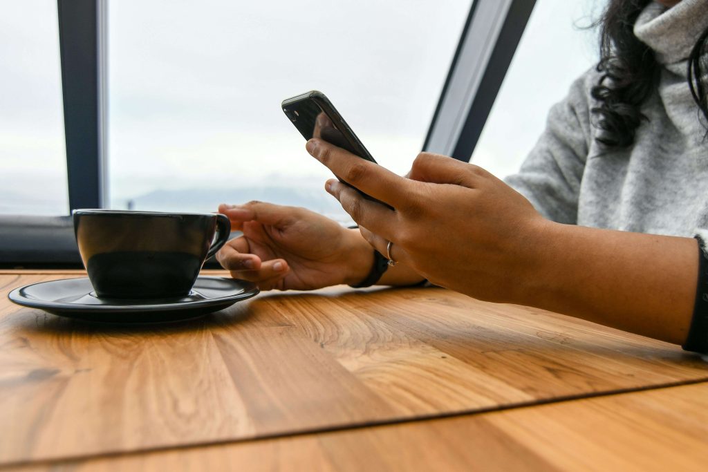 woman holding cell phone | eating disorder recovery coaching in the uk | eating disorder recovery coach | eating disorder coaching | meal support | England | Scotland | Ireland Image of a woman sitting at a table holding a cell phone while grabbing a coffee mug. Learn to maintain your healthy habits and continue to heal from your eating disorder with the help of a supportive eating disorder recovery coach in the UK.