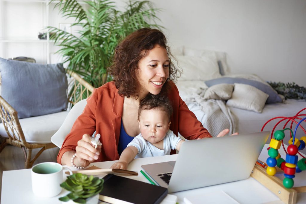 Mom sitting at a desk smiling and looking at a laptop while holding a baby. Find support during the postpartum period with your body image and stress with the help of an eating disorder recovery coach in England.