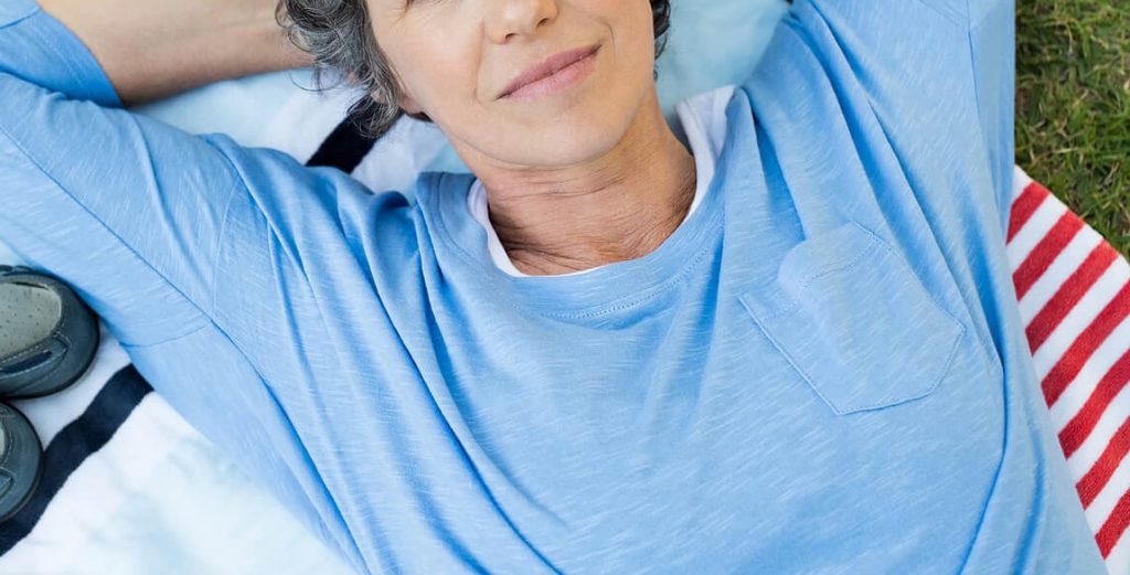 Thoughtful older woman laying on a blanket in the grass looking up at the sky with her hands behind her head. By working with a certified eating disorder recovery coach in the UK you can begin to eliminate your eating disorder symptoms in positive ways.