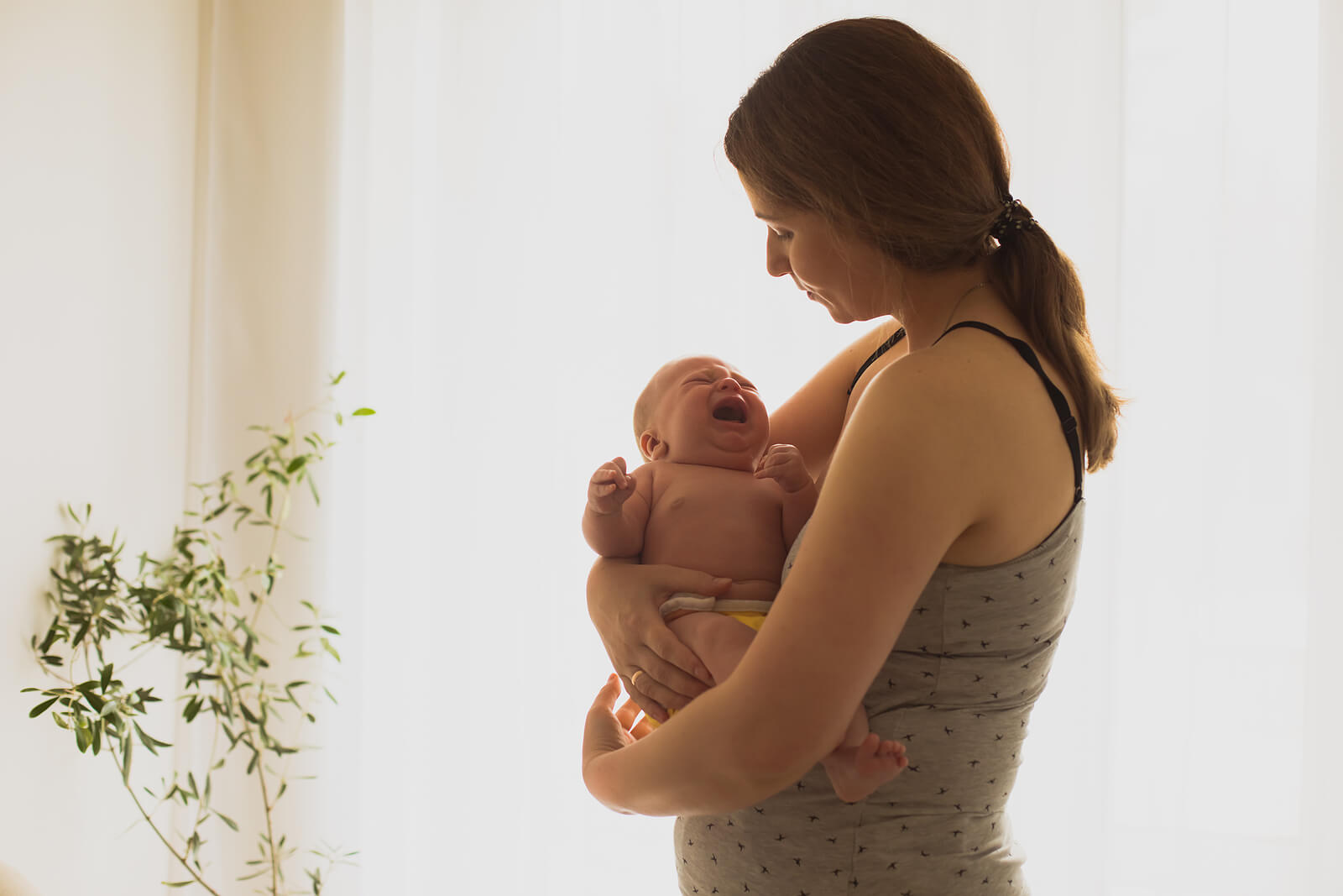 Postpartum mom standing by a window holding a newborn baby. If you're struggling with a postpartum eating disorder discover how compassionate eating disorder recovery coaching in England can help you manage your symptoms.