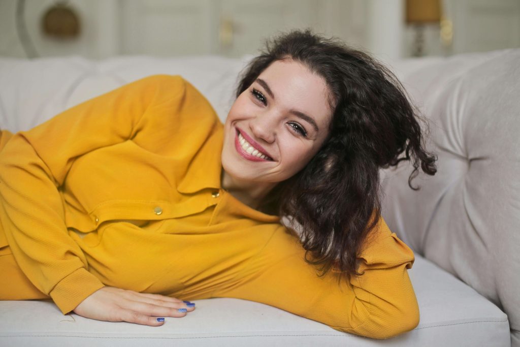 Smiling woman laying on a couch wearing a yellow shirt. Build confidence in yourself during eating disorder recovery with the help of eating disorder recovery coaching in the UK.