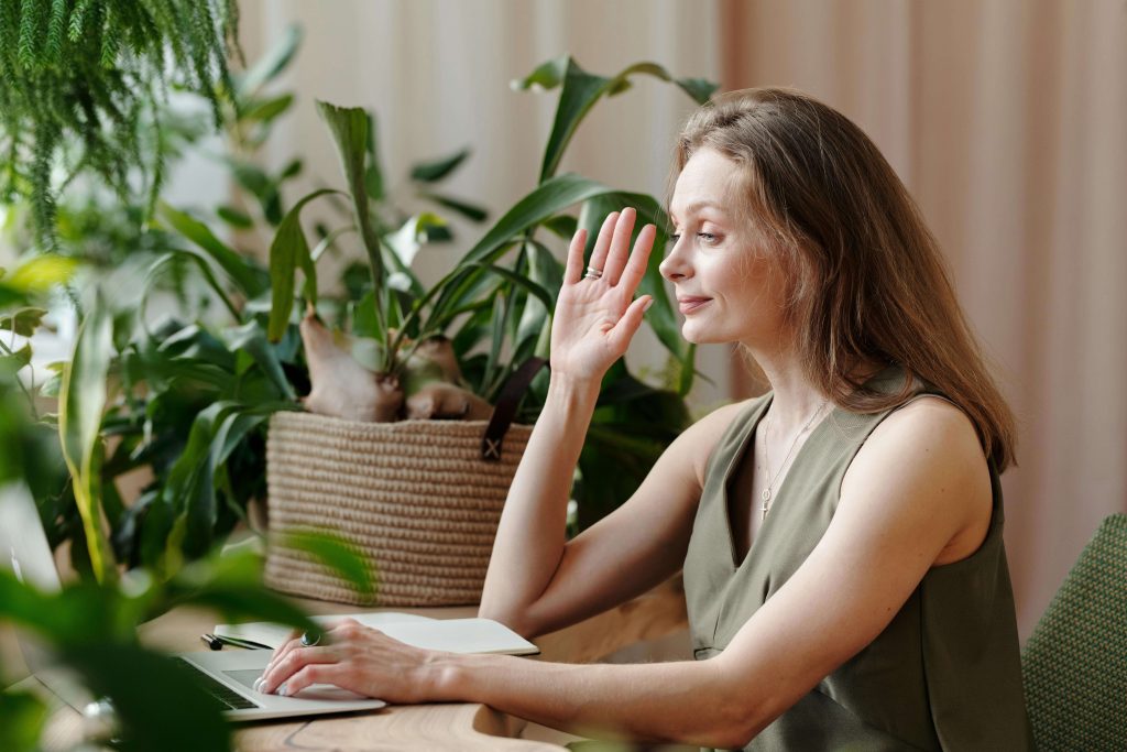 Smiling mature woman sitting at a table waving to a laptop. With the support of an eating disorder recovery coach in the UK, you can begin to overcome clean eating as a professional and manage unhealthy eating behaviors. 