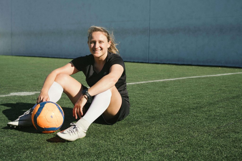 Smiling woman sitting on a football field with a football. Heal your relationship with food and exercise as an athlete with the support of eating disorder recovery coaching in the UK. 