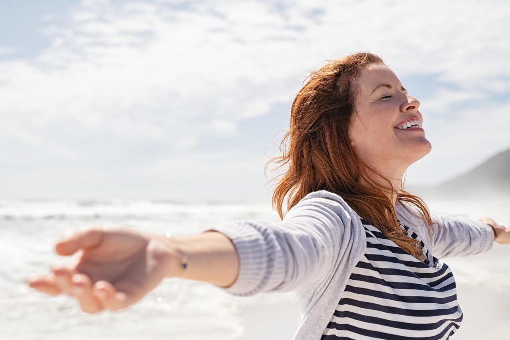 Happy redheaded woman standing on a beach with her eyes closed on a sunny day stretching her arms out. Heal your relationship with food and yourself with the help of eating disorder recovery coaching in England.
