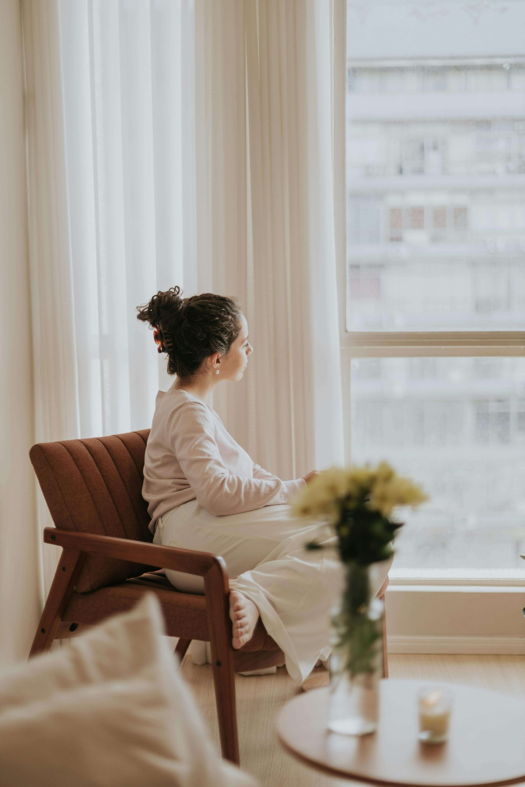 Thoughtful woman sitting in a chair next to a window looking out. Find support when it comes to managing and healing from extreme hunger with eating disorder recovery coaching in England.