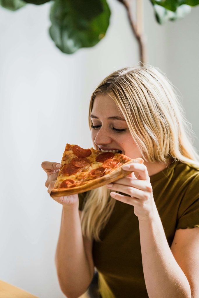 Woman sitting at a table taking a bite of pepperoni pizza. With the support of a British eating disorder recovery coach in the UK you can begin to manage your extreme hunger in healthy ways.