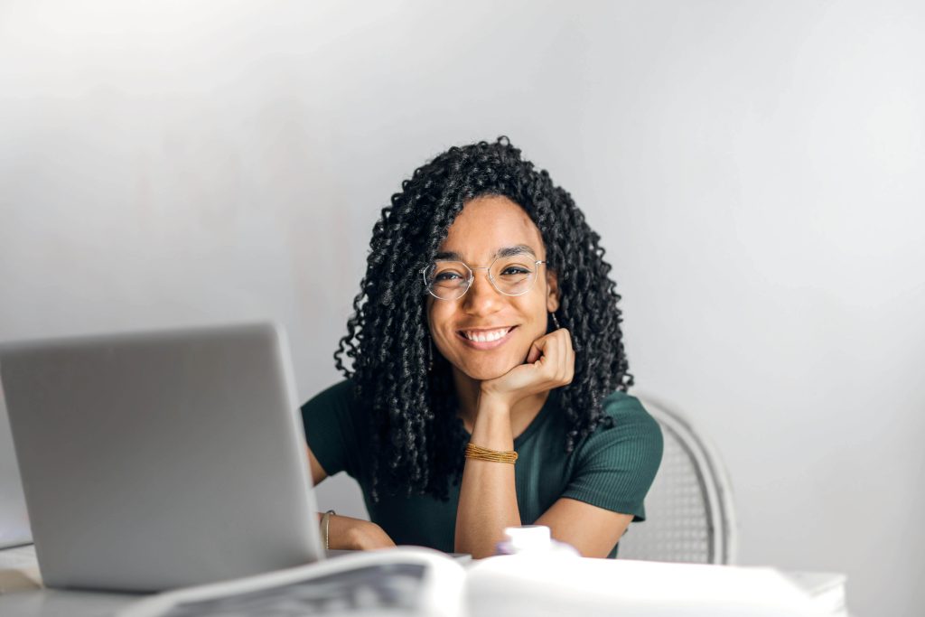 Smiling woman resting her chin on her hand while she sits at a desk with a laptop. With eating disorder recovery online in England, you can overcome your fear of food and heal in positive ways.