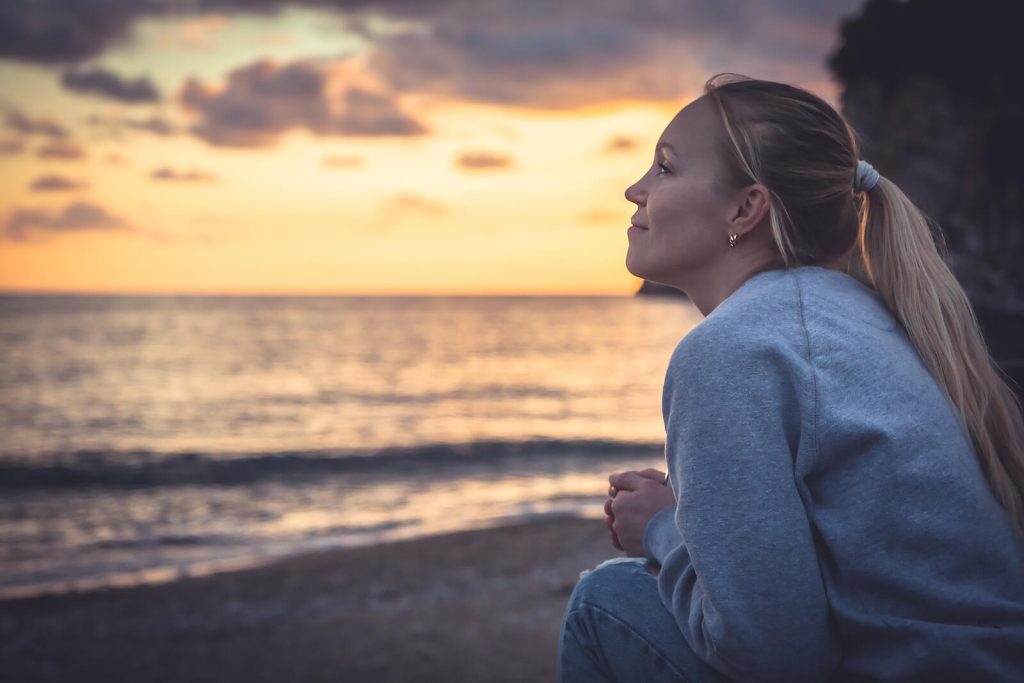 Woman sitting on the beach at sunset, gazing peacefully at the horizon. Eating disorder recovery online in England helps you restore self-compassion and rebuild trust with your body in England.