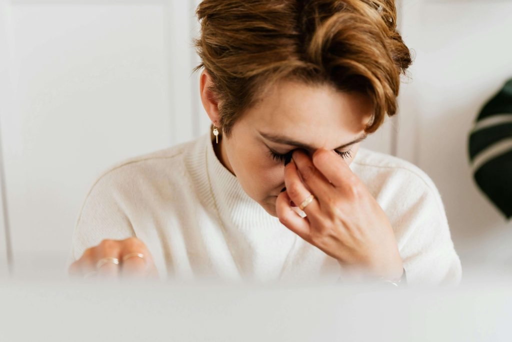 Stressed woman in a white sweater sitting indoors, rubbing her eyes in frustration. When stress and triggers feel overwhelming, eating disorder recovery coaching in England offers strategies to find relief and regain balance.