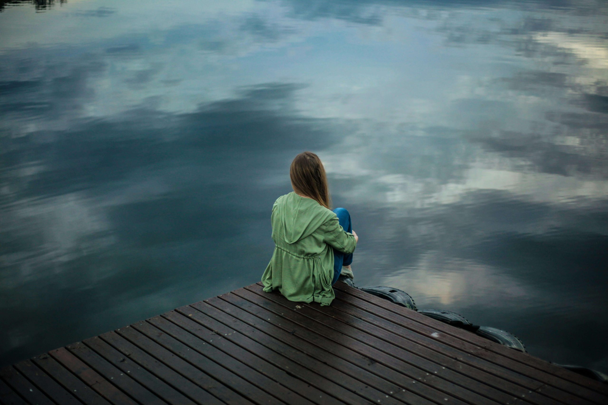 Woman in a green jacket sitting alone on a dock overlooking calm water, appearing contemplative. Overcome feelings of isolation with the support of an eating disorder recovery coach in England who can help you manage and move past triggers.
