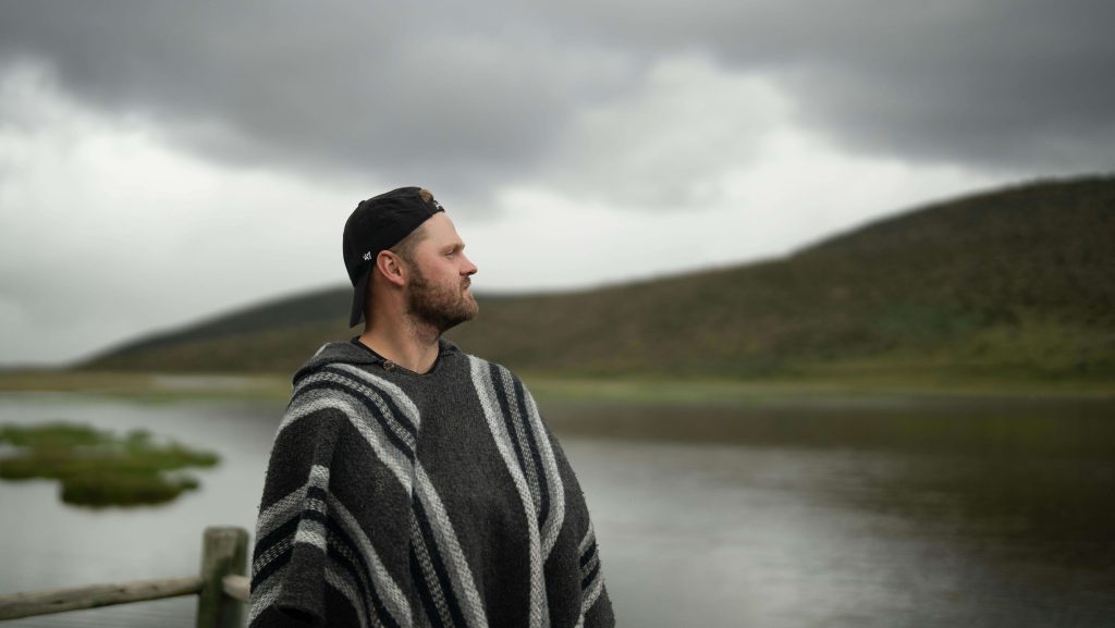 man near lake | eating disorder recovery coaching in the uk | British eating disorder recovery coach | eating disorder recovery online | England | Scotland | Ireland Man wearing a poncho and cap by a calm lake with cloudy skies. Work with a British eating disorder recovery coach to heal and rebuild confidence beyond the eating disorder.
