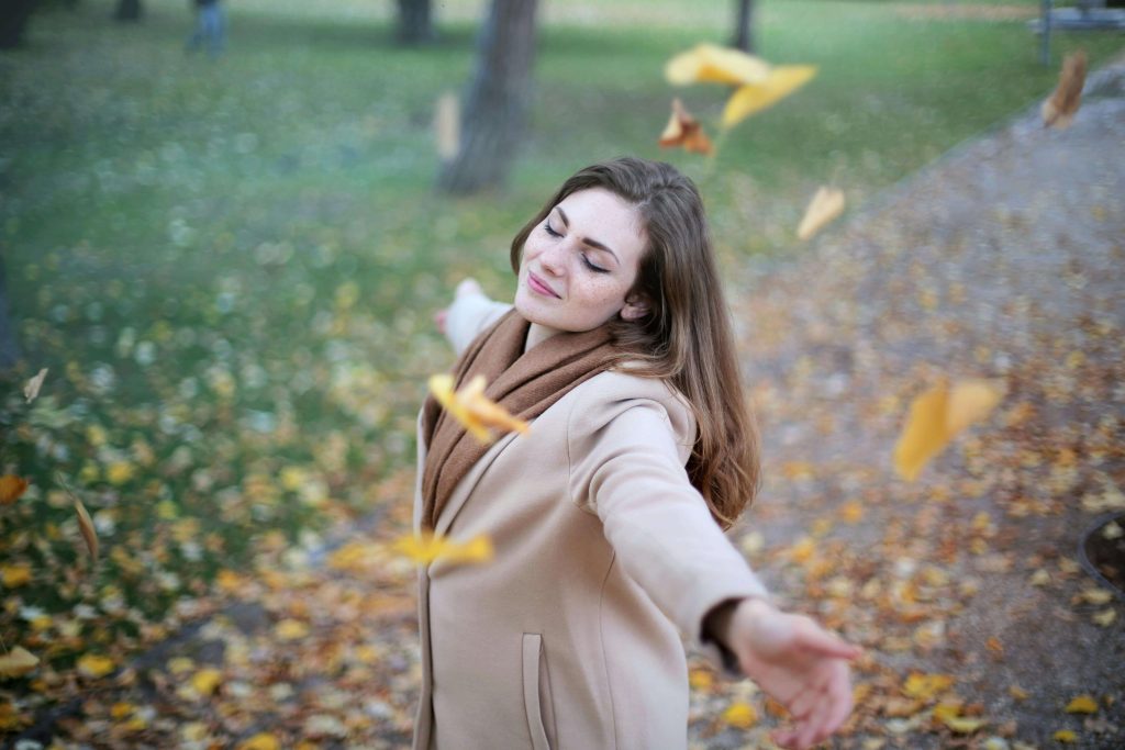 leaves | eating disorder recovery coaching in England | body checking | British eating disorder recovery coaching | England | Scotland | Ireland Woman standing in a park with arms outstretched, eyes closed, enjoying falling autumn leaves and a sense of freedom. Find guidance and face body checking with support to overcome your eating disorder with eating disorder recovery coaching in England.