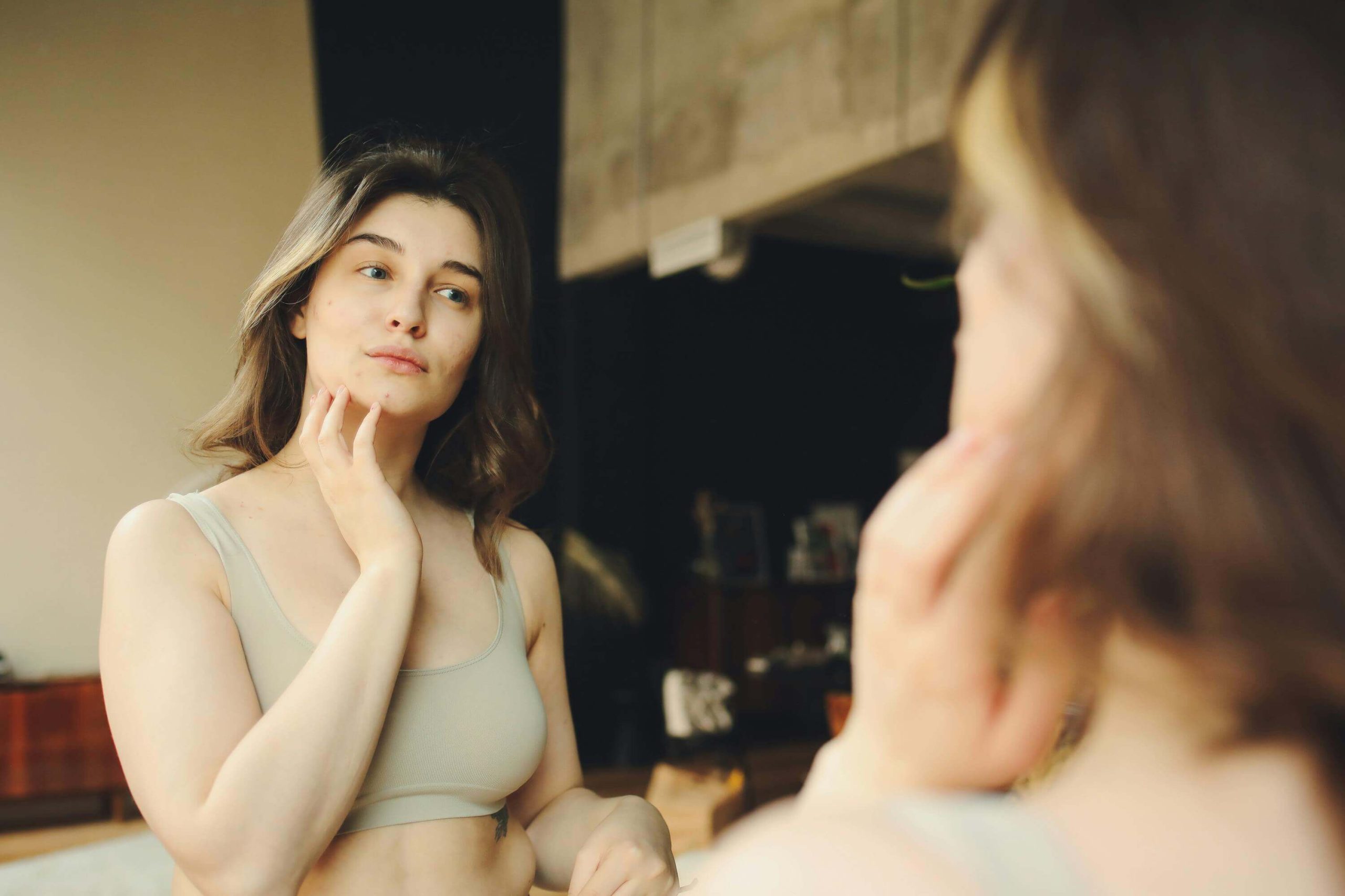 Woman looking at herself in the mirror and touching her face thoughtfully, noticing details in her reflection. With the support of an eating disorder recovery coach in England you can begin to overcome body checking in healthy ways.