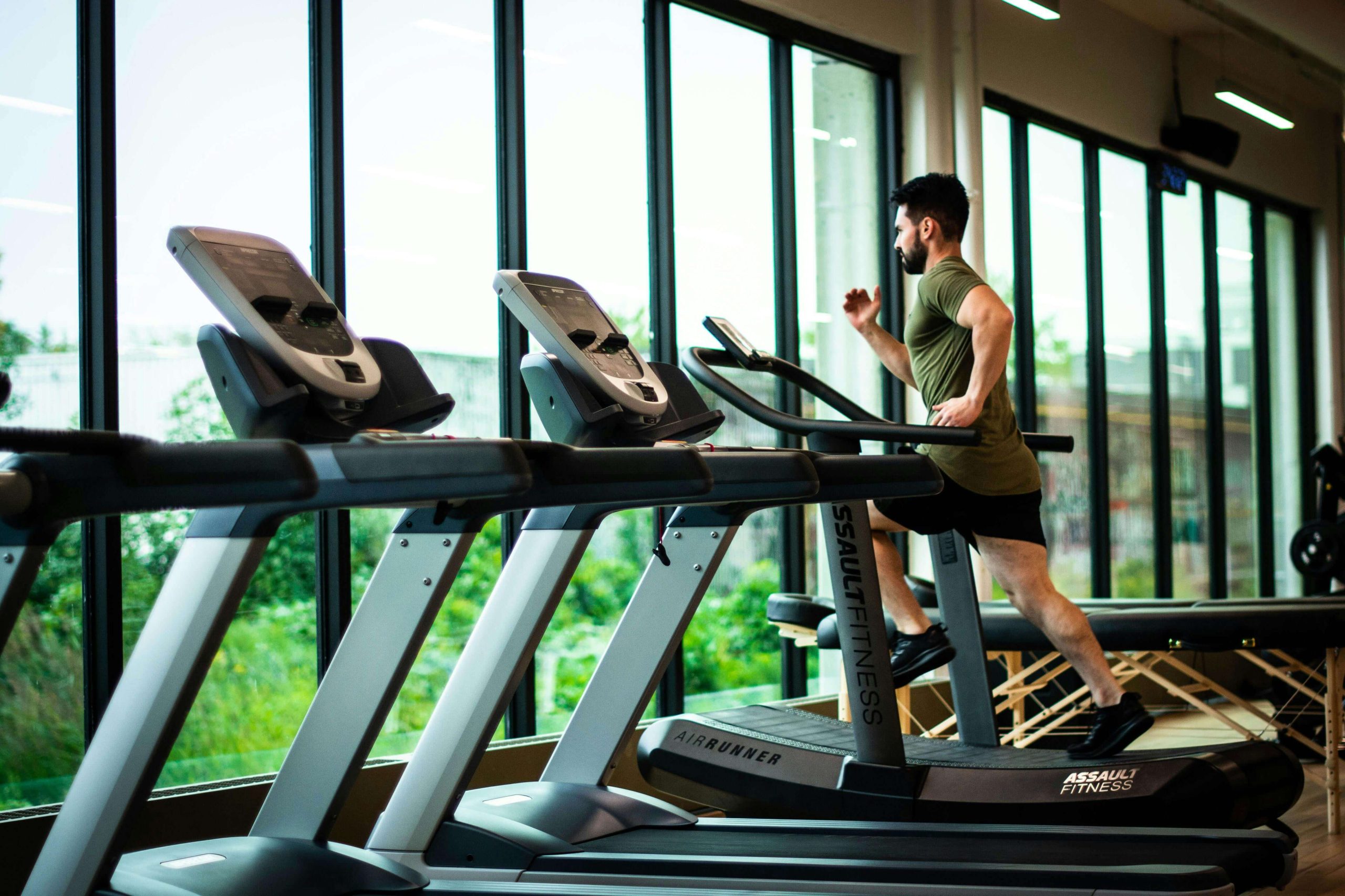 Man running on a treadmill in a modern gym with large windows overlooking greenery. Begin to cope with your exercise addiction and start balancing your life in healthy ways with eating disorder recovery coaching in England.