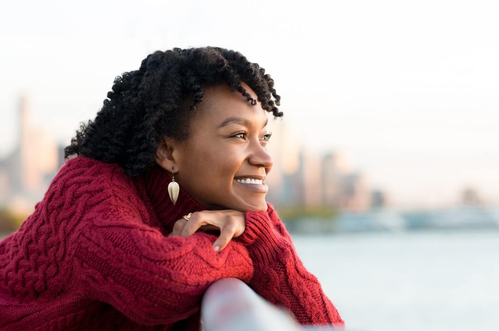 Young woman in a red sweater smiling while leaning on a railing with a city skyline in the background. Find steady, compassionate support at every stage with anorexia recovery coaching in England.