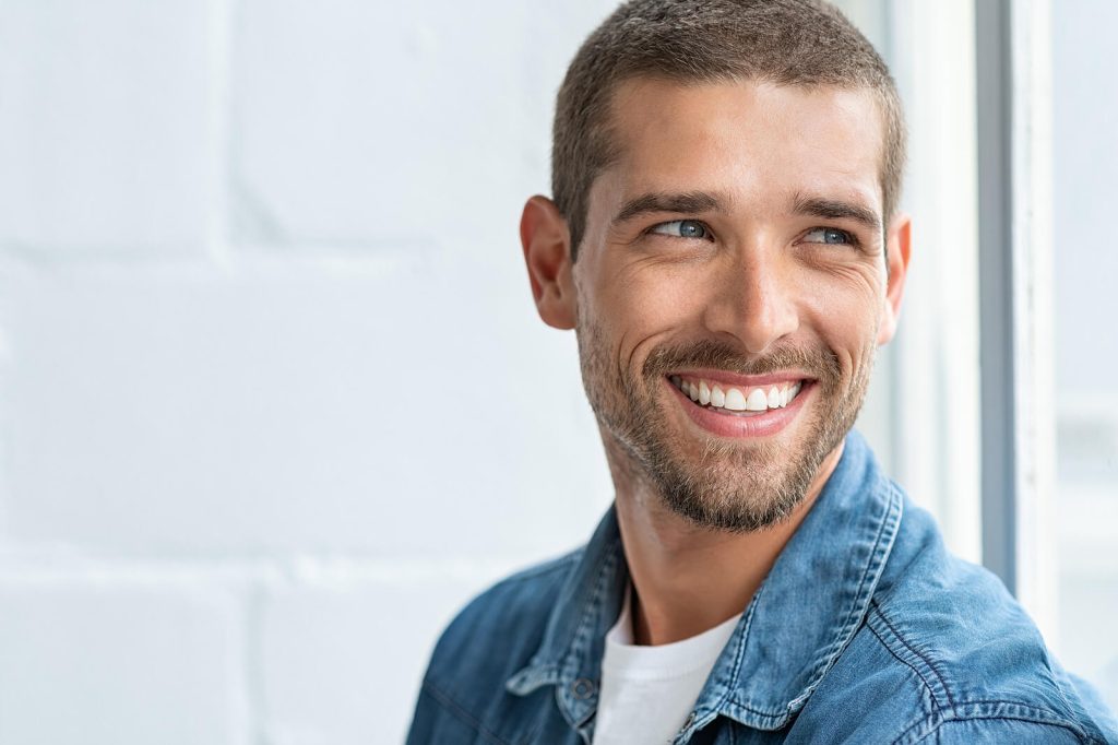 Smiling man wearing a denim shirt, standing indoors near a window with natural light. Move toward freedom and a calmer relationship with food through anorexia recovery coaching in England.