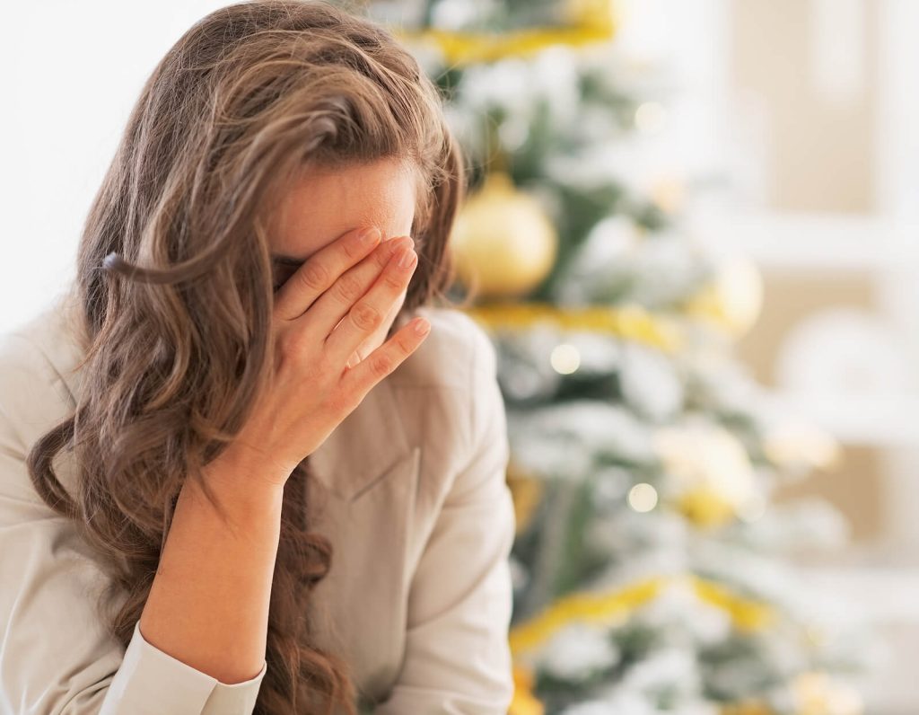 upset woman | anorexia recovery coaching in england | eating disorder recovery coach | anorexia nervosa recovery | London | Birmingham | Bath A woman sitting near a Christmas tree with her head in her hands, appearing overwhelmed or distressed during the holiday season. Begin finding steadier support during the festive season through anorexia recovery coaching in England to ease moments that feel emotionally heavy.