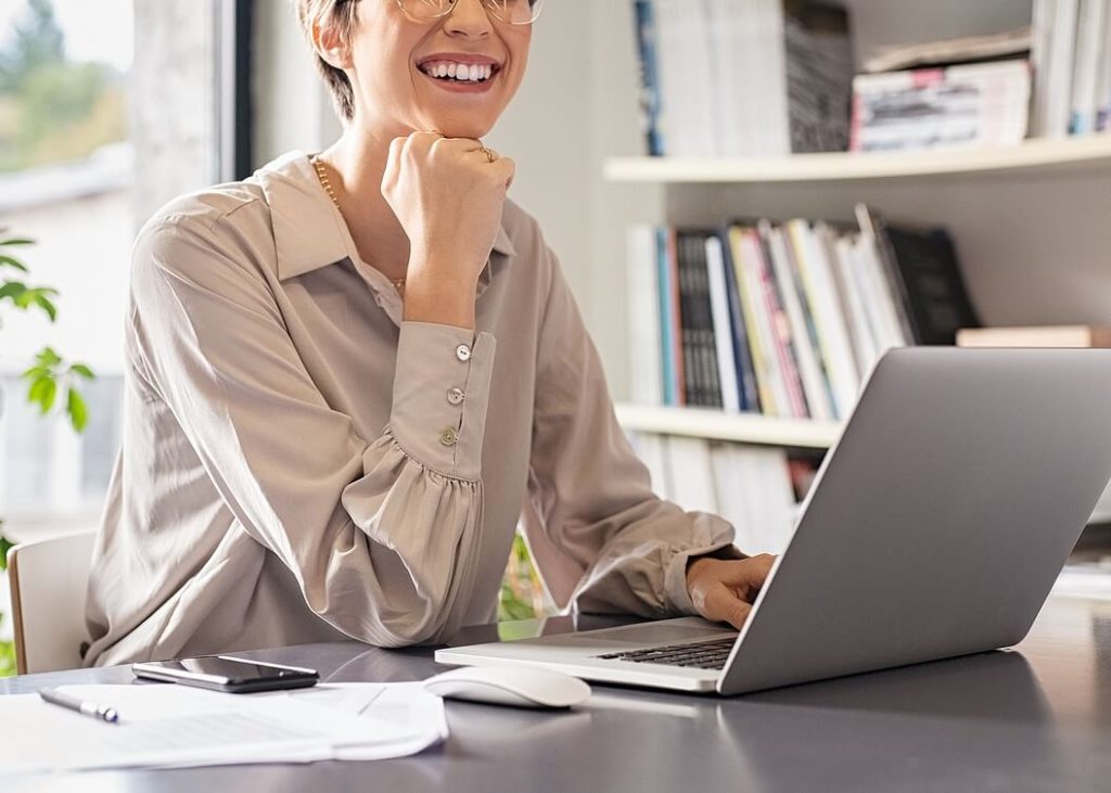 Woman with glasses sitting at a desk, smiling while working on a laptop in a home office. Take meaningful steps toward recovery and self-trust with anorexia recovery coaching in England.