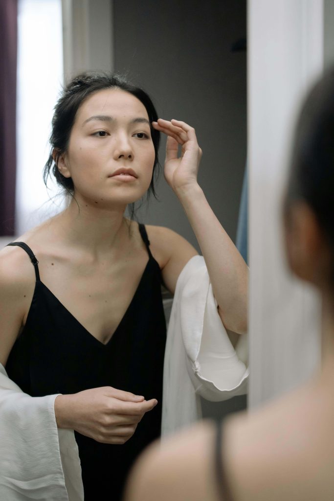 A woman gently touches her face while looking into a mirror, reflecting a moment of quiet self-examination. Find clarity beyond the eating disorder with anorexia recovery coaching in England, helping you reconnect with who you are beneath the rules and restrictions.