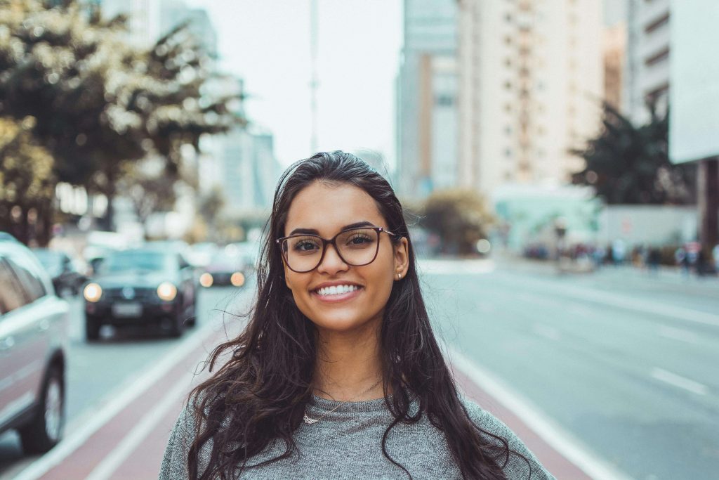 Young woman with long dark hair and glasses smiling while standing on a busy city street. An anorexia coach offering anorexia recovery coaching in England can help you begin your anorexia nervosa recovery with confidence.