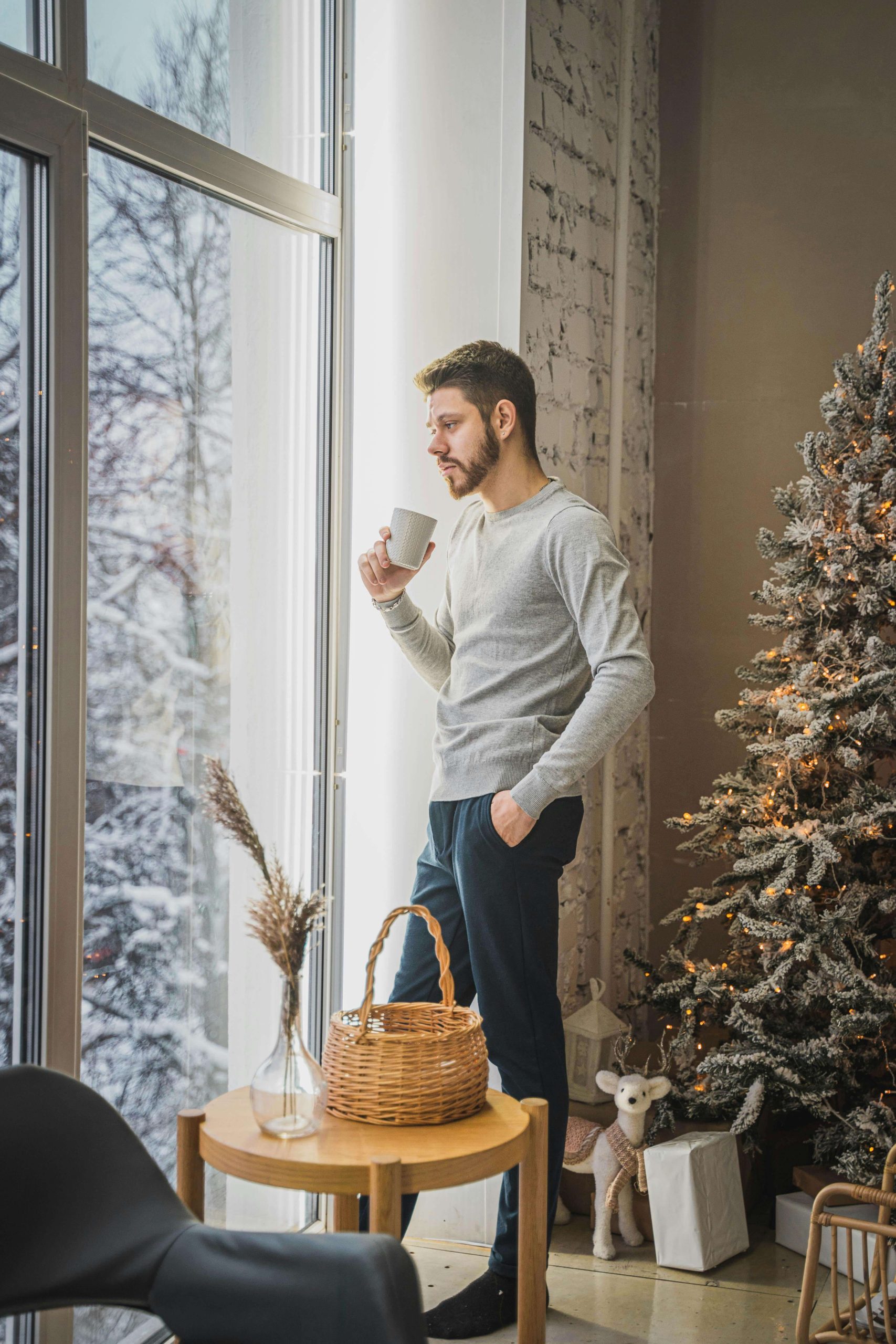 A man standing by a large window holding a mug, gazing outside. Find space for reflection and healing this winter season through personalised anorexia recovery coaching in England guided by an experienced eating disorder recovery coach.