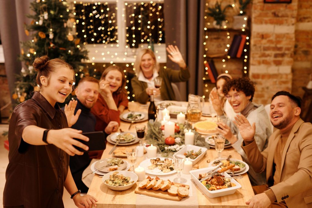 friends | anorexia recovery coaching in england | eating disorder recovery coach | anorexia nervosa recovery | London | Birmingham | Bath A family gathered around a festive Christmas dinner table, smiling and waving as someone takes a group selfie. Navigate holiday meals with more confidence by working with an eating disorder recovery coach who offers anorexia recovery coaching in England.