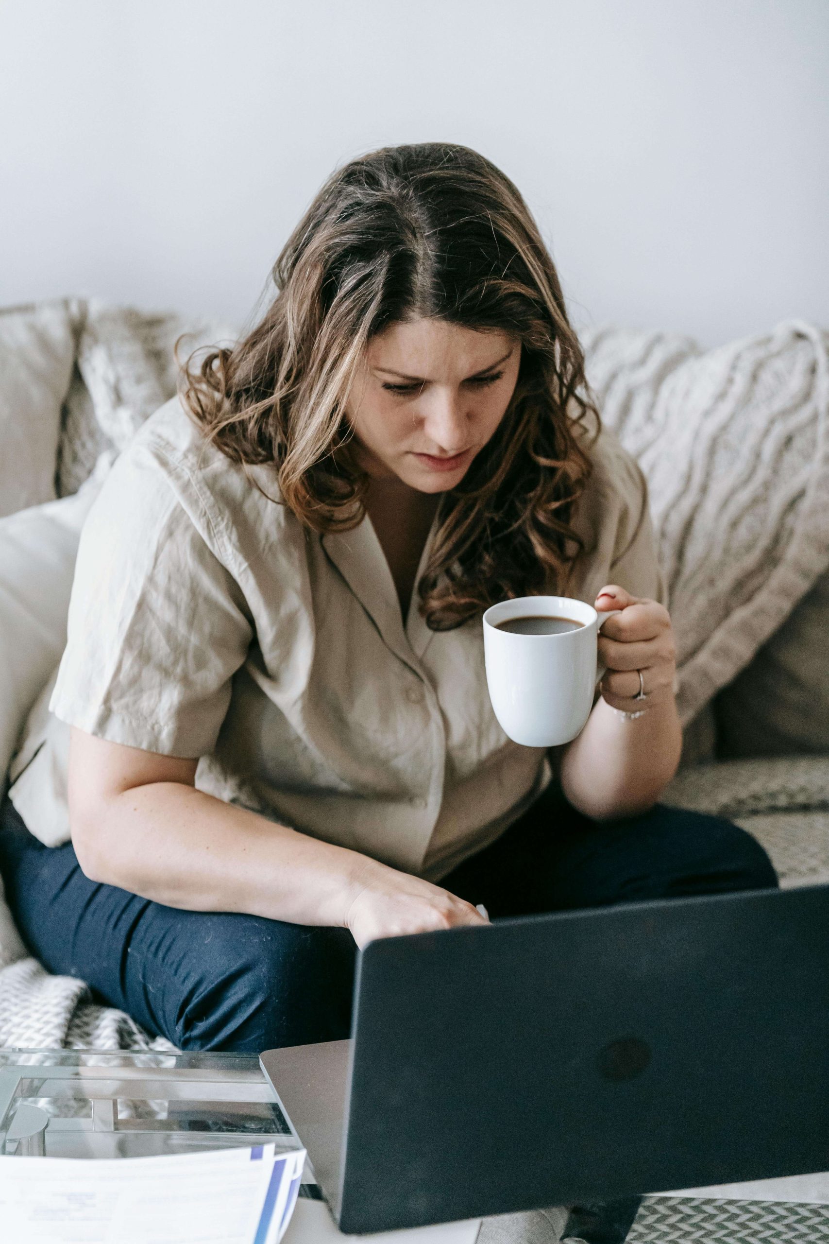 Woman sitting on a couch holding a coffee mug while working on a laptop at home. Through online anorexia recovery coaching in England, an eating disorder recovery coach can guide you as you rebuild strength in your anorexia nervosa recovery.