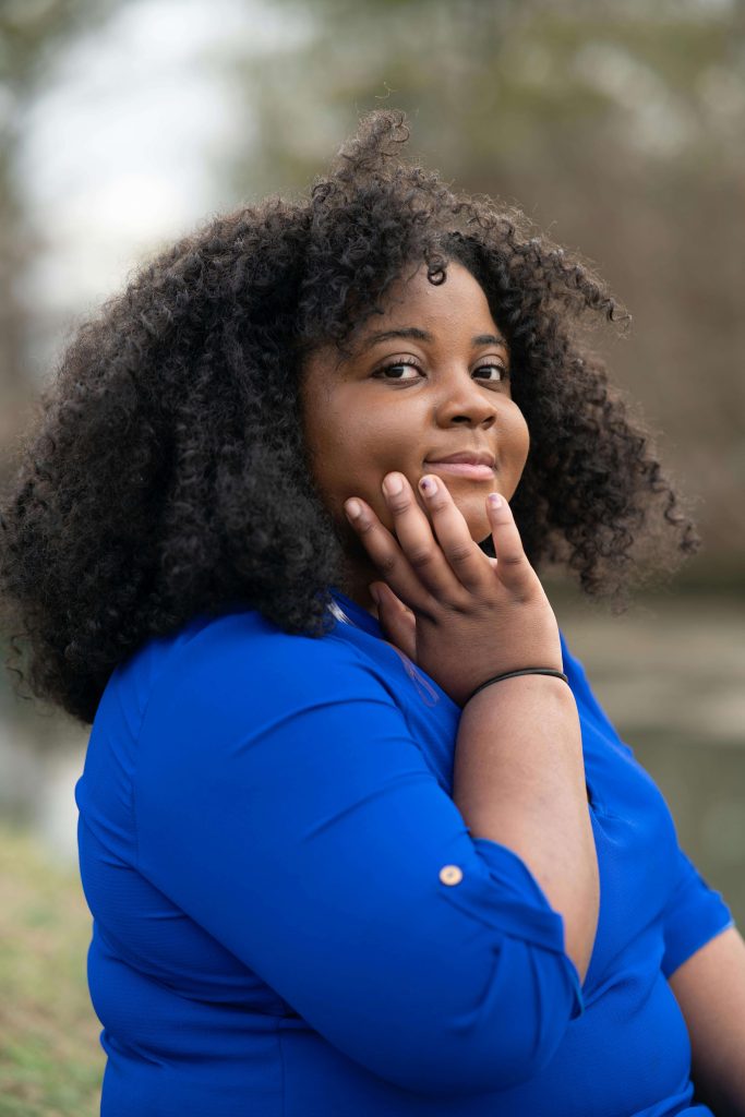 Confident woman with curly hair wearing a bright blue shirt, resting her hand on her cheek while posing outdoors. With anorexia recovery coaching in England, an anorexia coach can help you take encouraging steps toward your anorexia nervosa recovery.