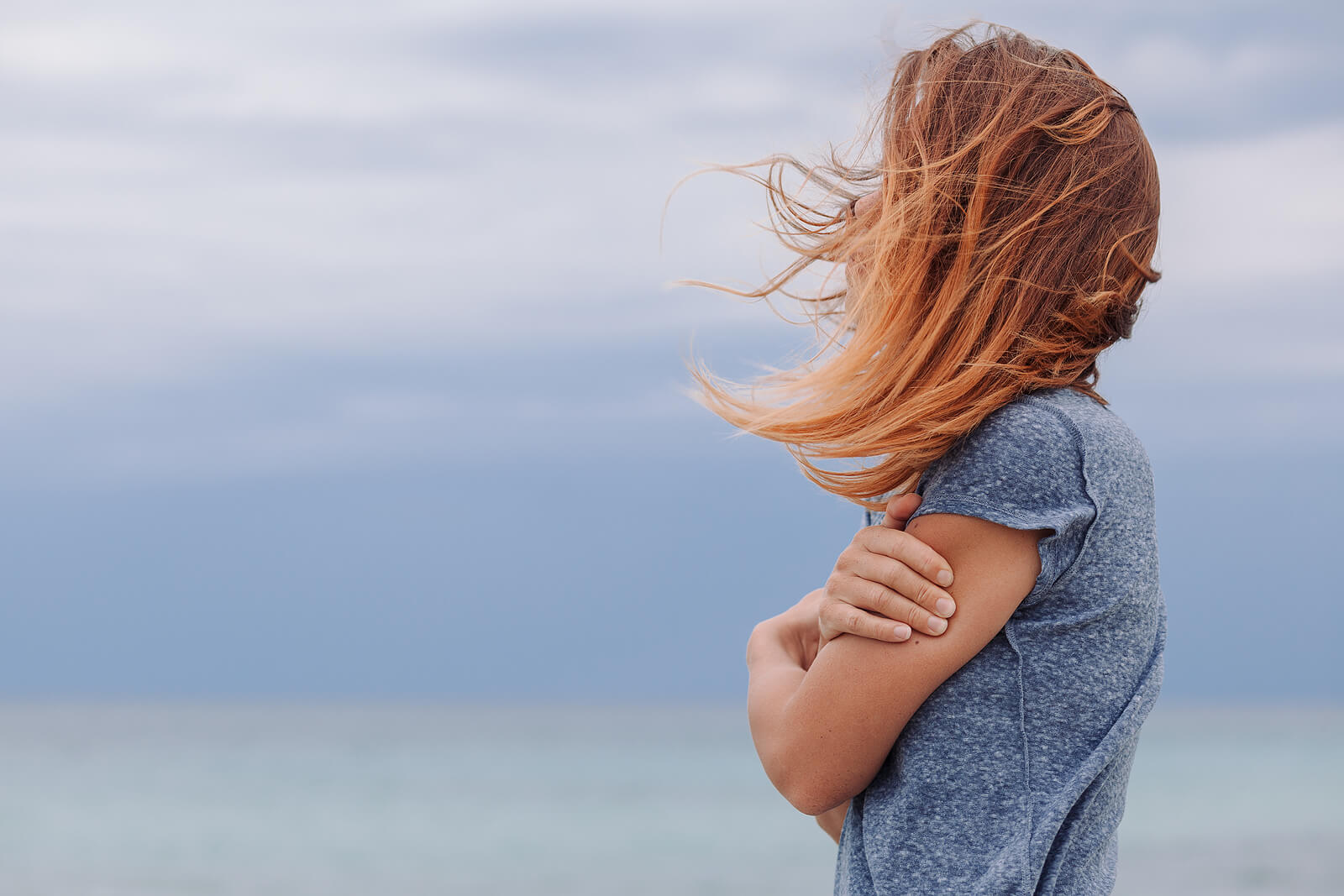 Person standing outdoors near the sea with arms crossed, hair blowing in the wind, looking toward the horizon. Find steady, compassionate support for food-related guilt through eating disorder recovery coaching in England.