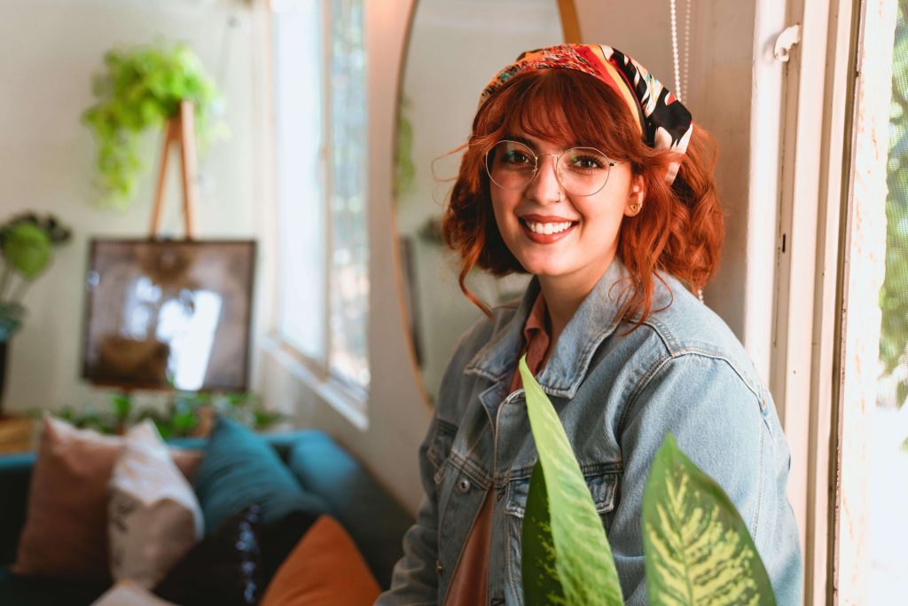 Smiling person with red hair and glasses standing indoors near a window, surrounded by houseplants. Move toward a calmer, more confident relationship with food with eating disorder recovery coaching in England.