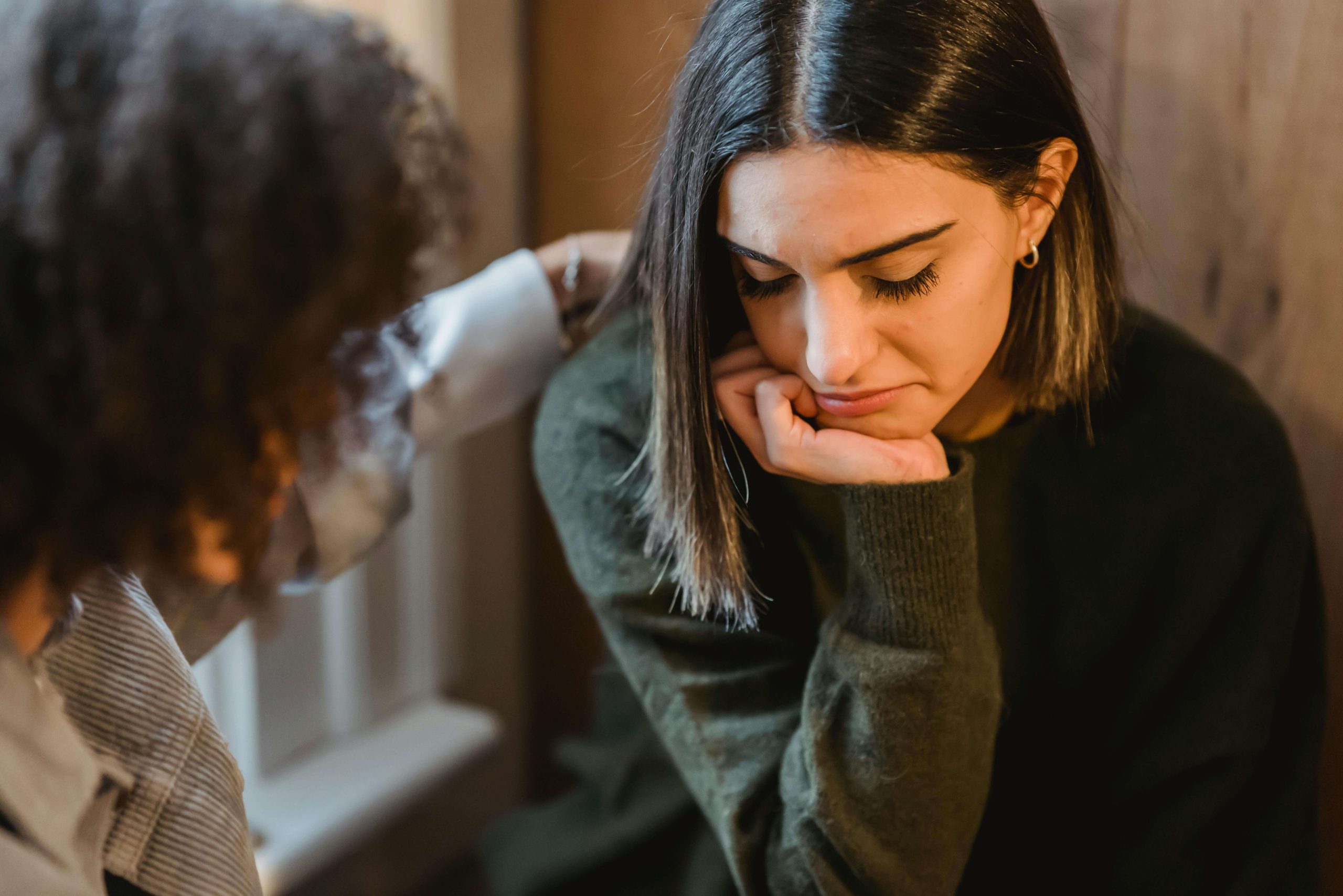 Worried woman in green sweater with hand on chin while someone comforts her. Find support for caretaker fatigue through eating disorder recovery coaching in England.