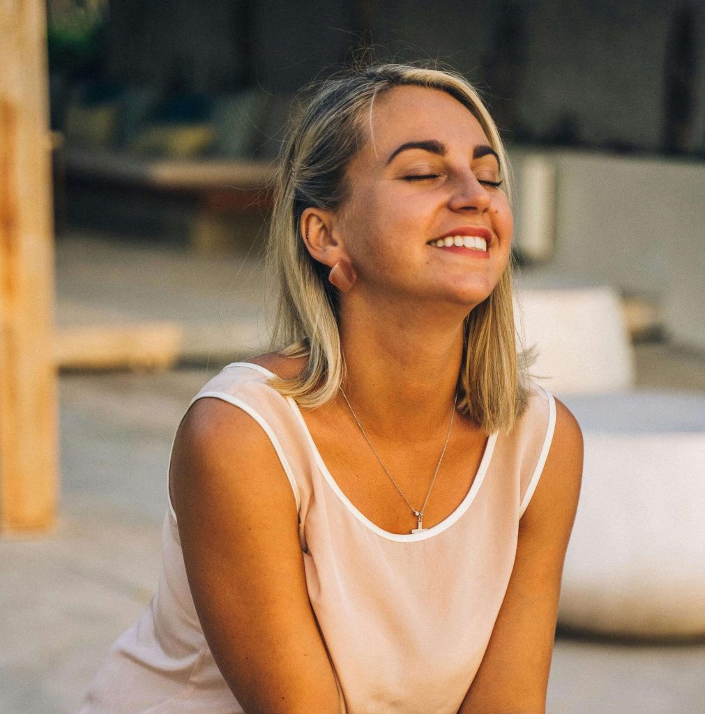 Woman smiling peacefully outdoors with eyes closed, holding a journal and pen. Create the consistency that leads to freedom with eating disorder recovery coaching in England.