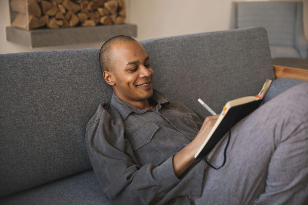 Man relaxing on gray couch while writing in a journal and smiling. Take small, daily steps toward lasting change through eating disorder recovery coaching in England.