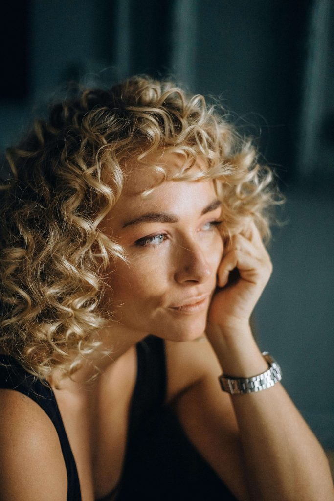 Woman with curly blonde hair resting her head on her hand in contemplation. Stop waiting for motivation and start building recovery habits with eating disorder recovery coaching in England.