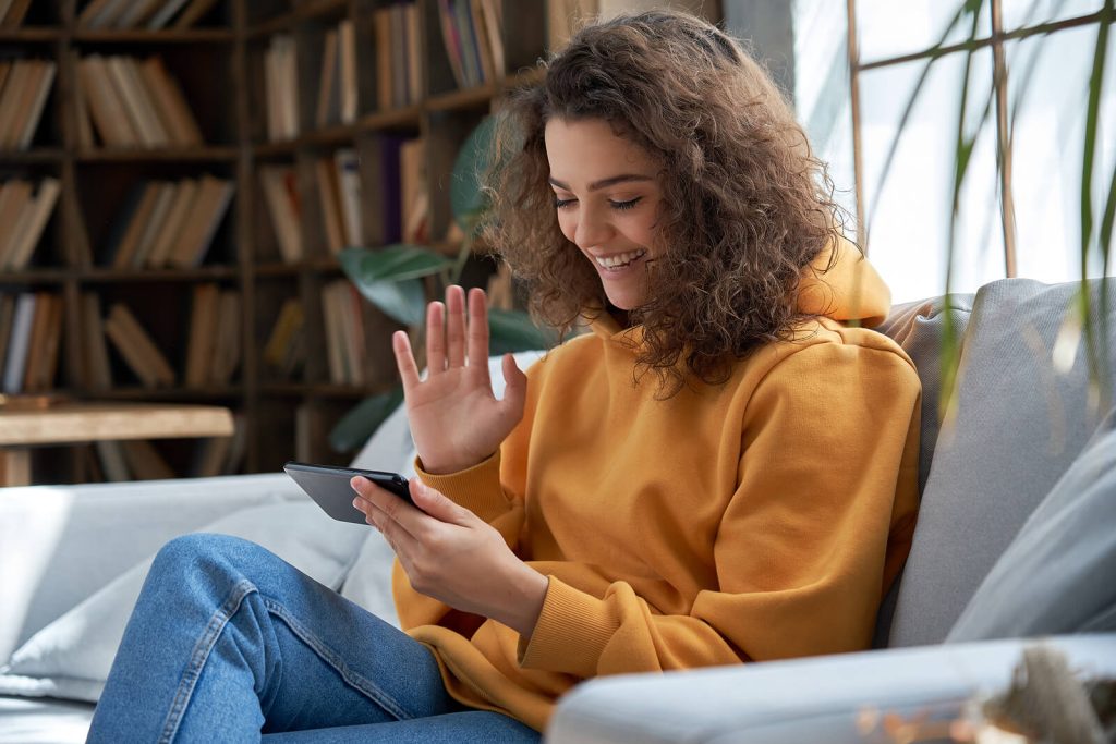 A young woman with curly hair sitting on a sofa in a yellow hoodie, smiling and waving at her phone during a video call. Access eating disorder recovery coaching in England online from wherever you are and take the first step towards freedom from food anxiety.