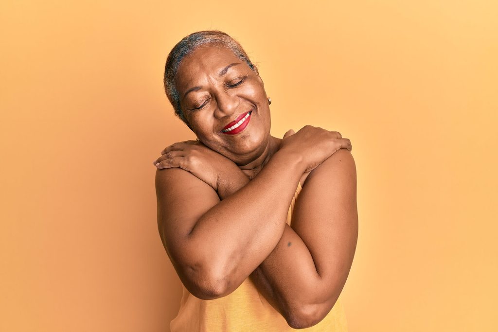 A older woman with short grey hair smiling warmly with her eyes closed, hugging herself against an orange background. Develop a kinder, more accepting relationship with your body with the support of eating disorder recovery coaching in England.