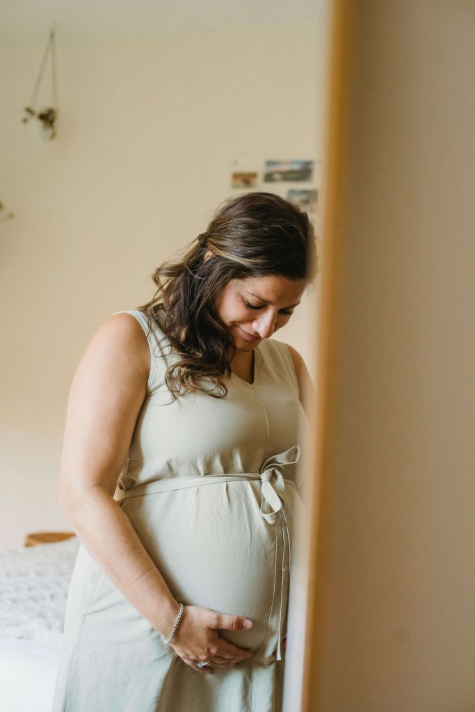 A pregnant woman with long dark hair standing in a softly lit room, looking down and cradling her bump in a sage green dress. Take the first step towards a calmer pregnancy with compassionate eating disorder recovery coaching in England by your side.