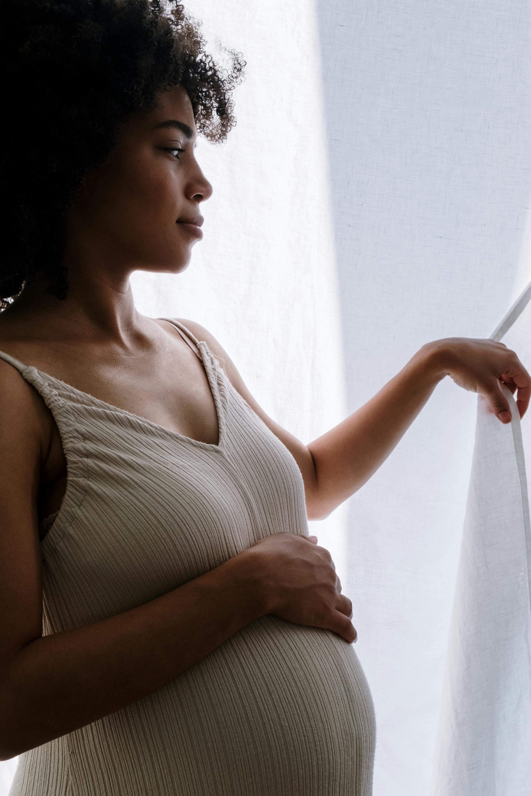 A pregnant woman with curly hair standing by a window, gazing thoughtfully to the side while gently holding her bump. You don't have to face eating disorder thoughts alone — eating disorder recovery coaching in England can help you find peace during pregnancy.