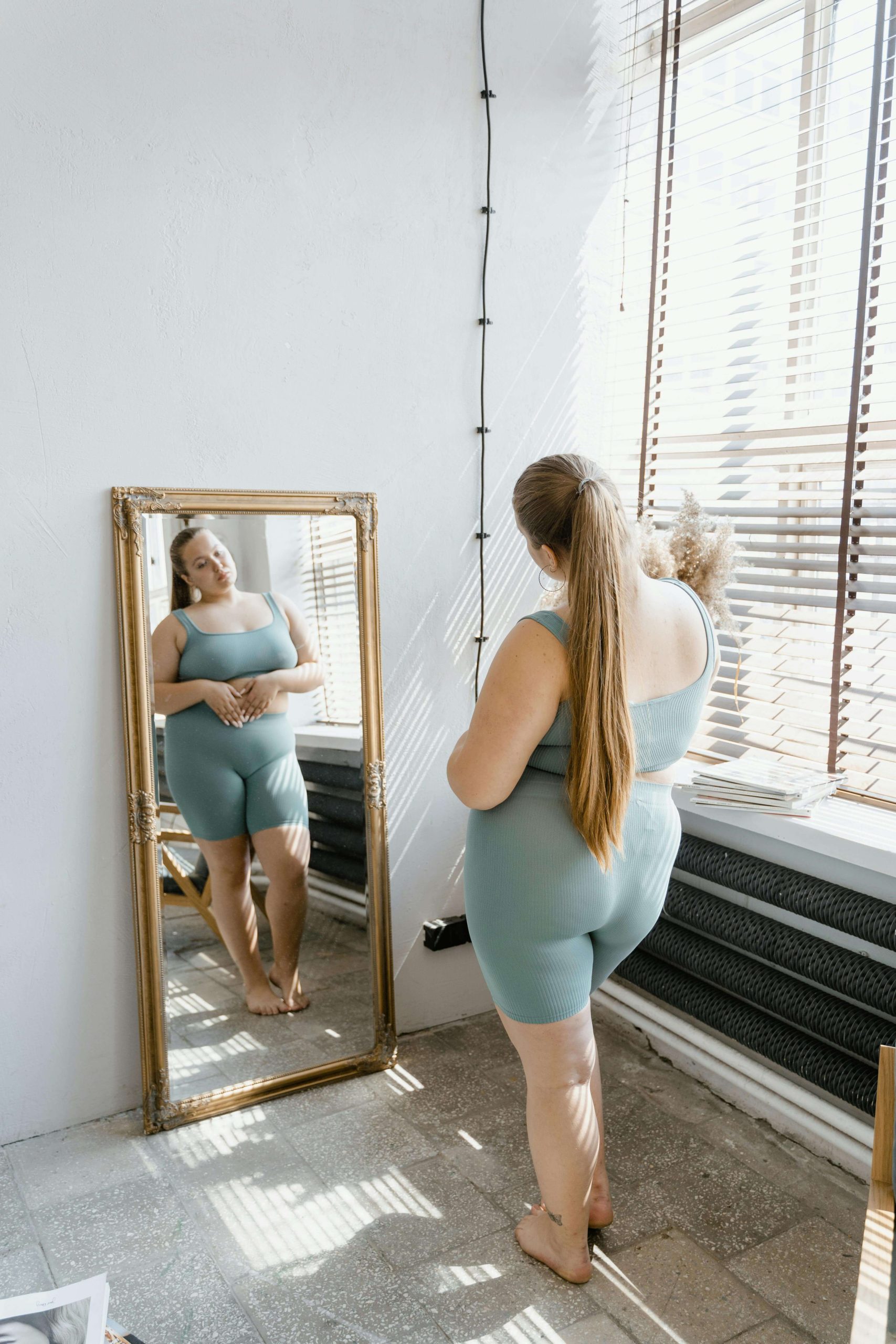 A woman in a blue co-ord set standing barefoot in front of a gold-framed full-length mirror in a bright room, looking at her reflection. Break free from body checking and learn to rebuild trust with your body through eating disorder recovery coaching in England.