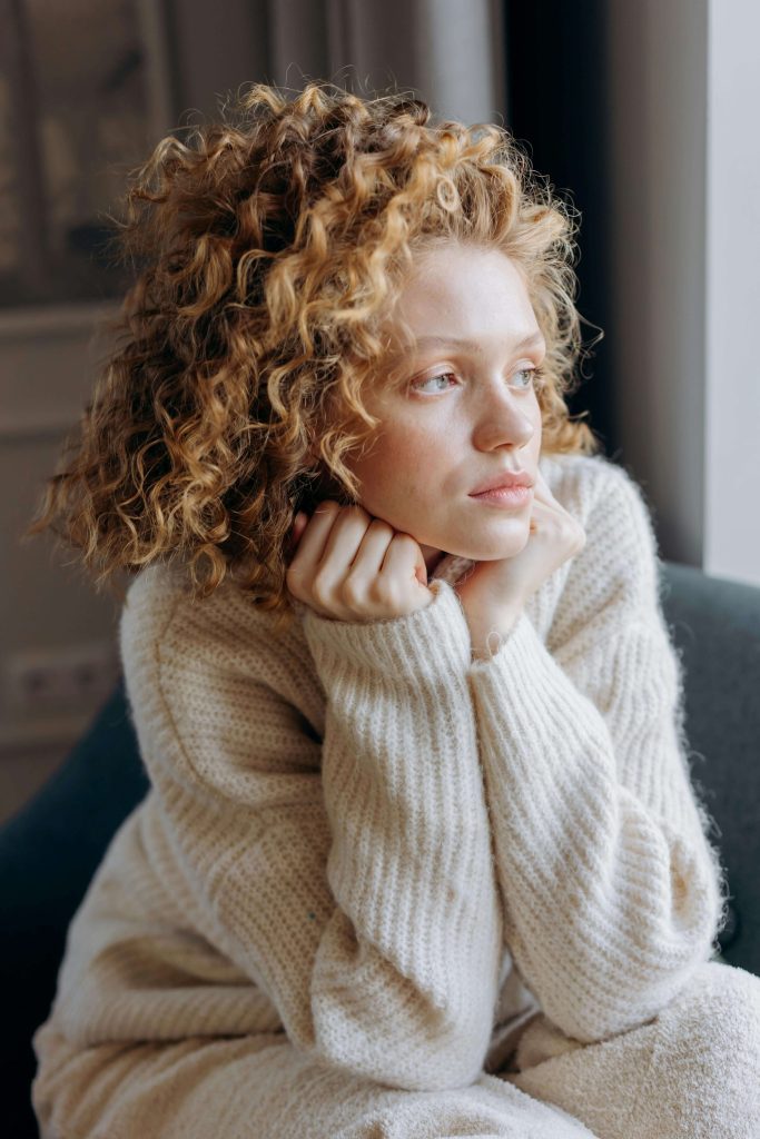 A young woman with curly blonde hair sitting in a cream knit jumper, resting her chin on her hands and gazing thoughtfully towards a window. If anxiety around food feels overwhelming and impossible to overcome, eating disorder recovery coaching in England can help you find a way forward.