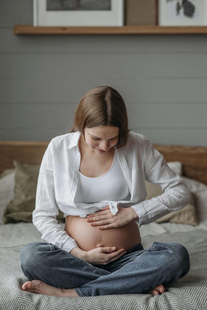 A pregnant woman with brown hair sitting cross-legged on a bed, looking down and cradling her bare bump with both hands. Find the support you deserve through eating disorder recovery coaching in England as you navigate the emotional challenges of pregnancy.